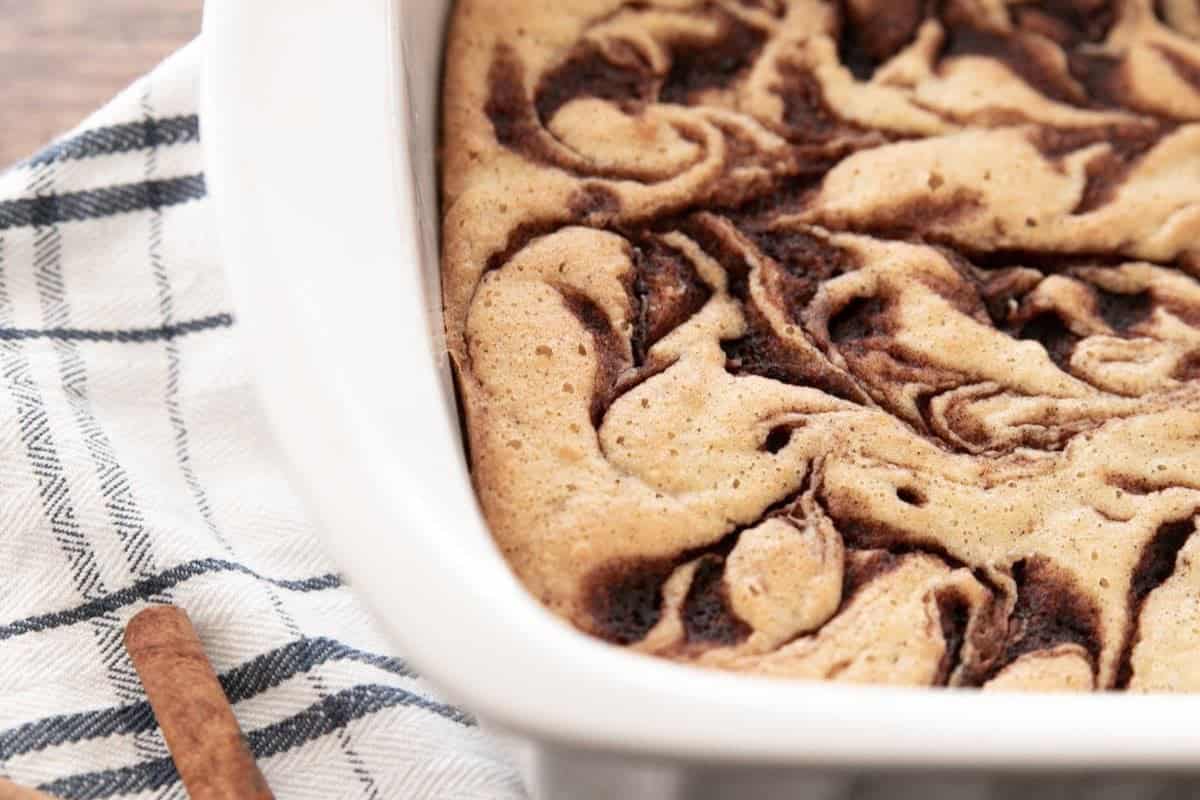 A close-up of a cinnamon swirl cake in a white baking dish, resting on a plaid kitchen towel, with part of a cinnamon stick visible nearby. The cake has a golden-brown top with dark cinnamon swirls.