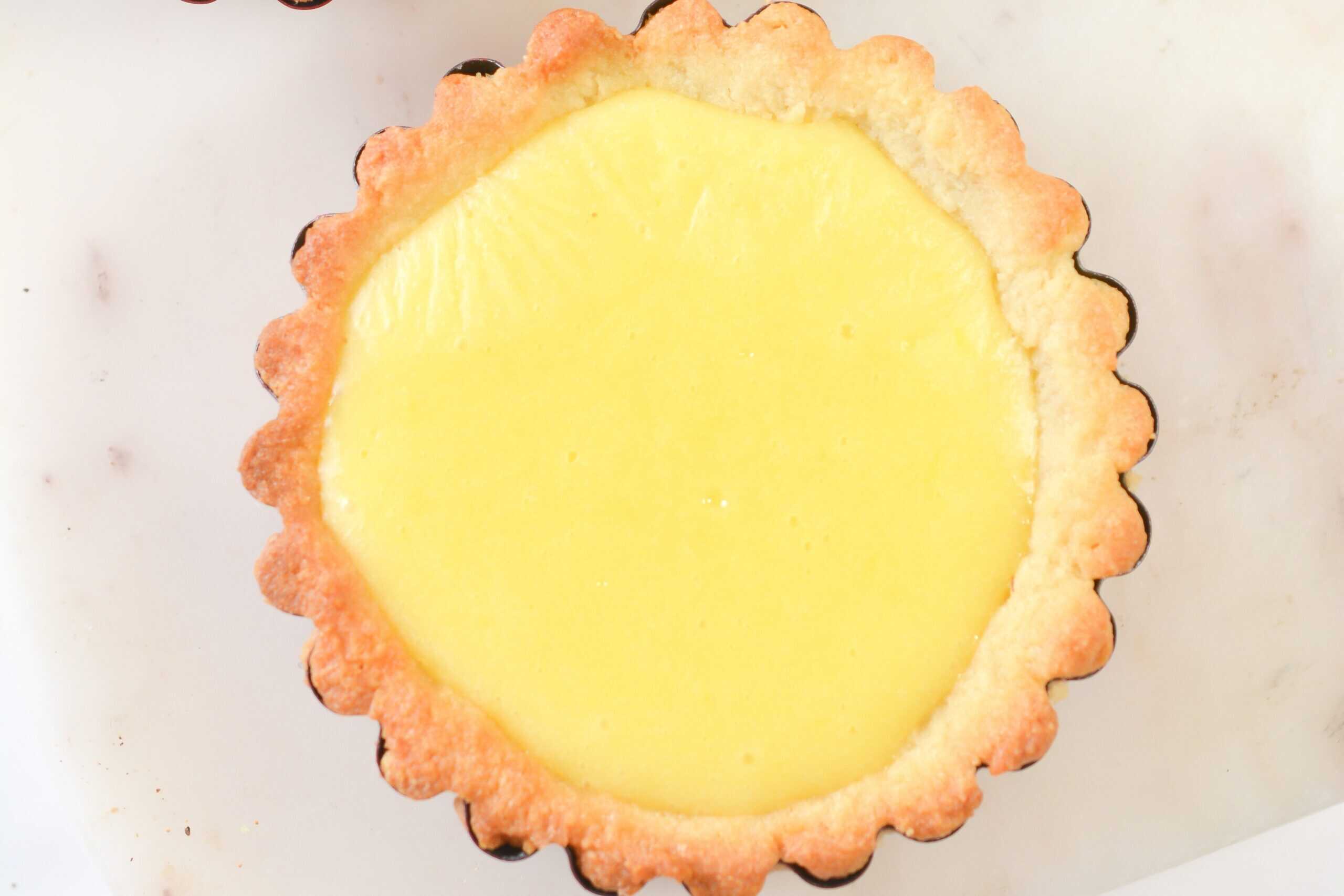 A small tart with a scalloped, golden-brown crust filled with smooth, yellow custard, viewed from above on a white surface.