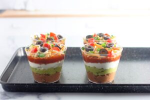 Four clear cups on a black tray, each filled with layered dip including refried beans, guacamole, sour cream, salsa, shredded cheese, diced tomatoes, sliced black olives, and chopped green onions.
