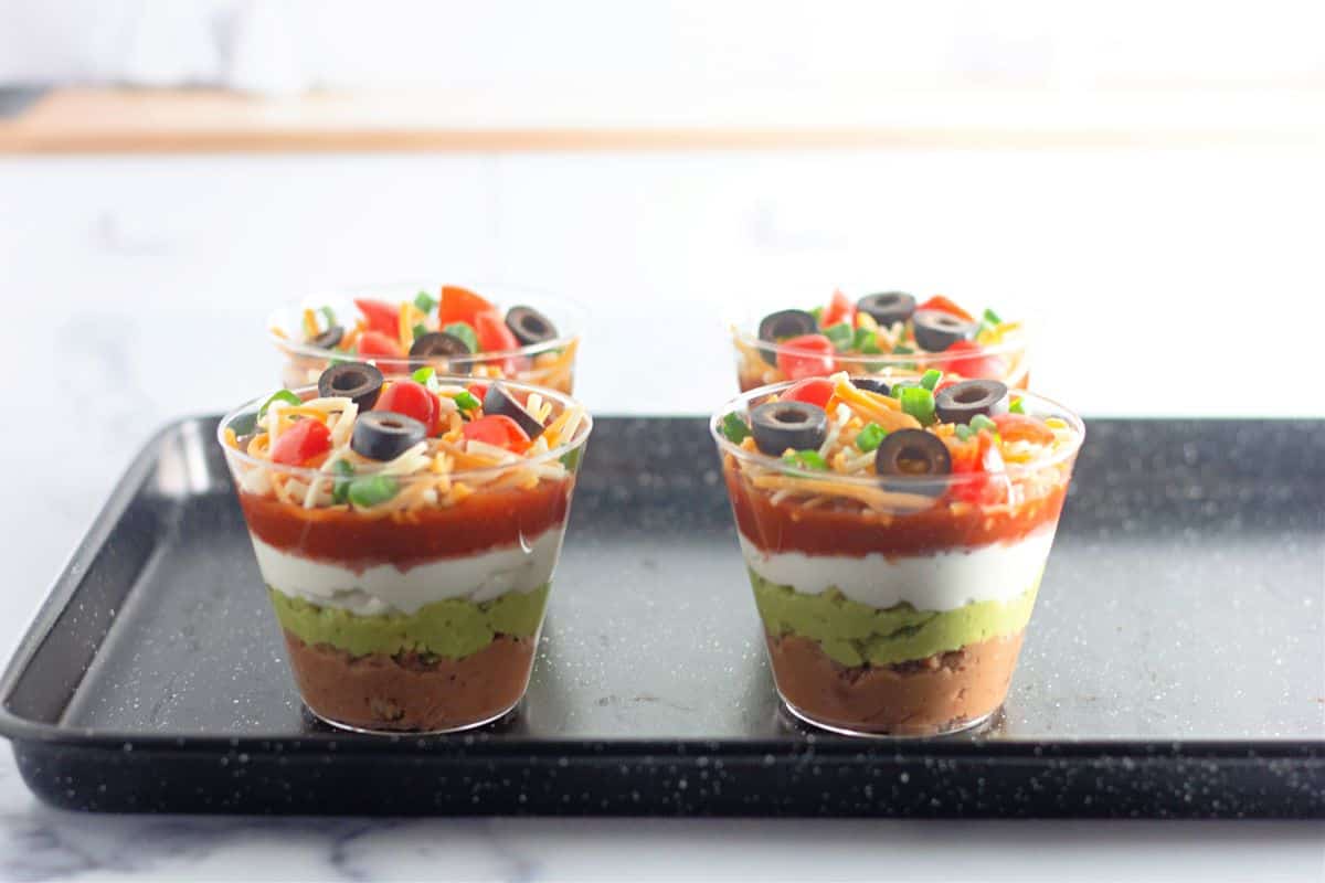 Four clear cups on a black tray, each filled with layered dip including refried beans, guacamole, sour cream, salsa, shredded cheese, diced tomatoes, sliced black olives, and chopped green onions.