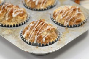 Muffins topped with crumbly streusel and white icing drizzle sit in a metal muffin tin lined with paper cups.