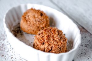 Two scoops of crumbly, brown cookies or cookie dough sit in a white, scalloped dish on a light wooden surface.