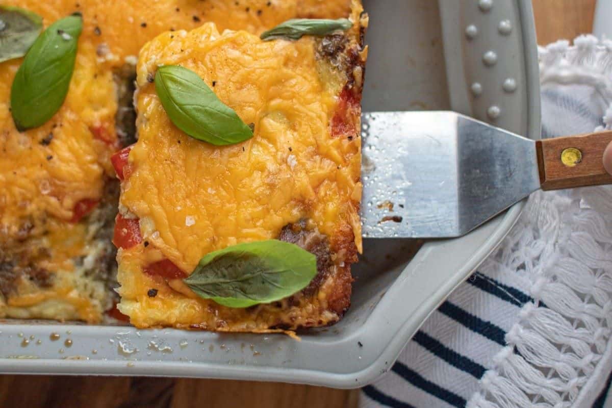 A spatula lifting a cheesy, baked casserole topped with fresh basil leaves from a gray baking dish, with a striped towel beside it.