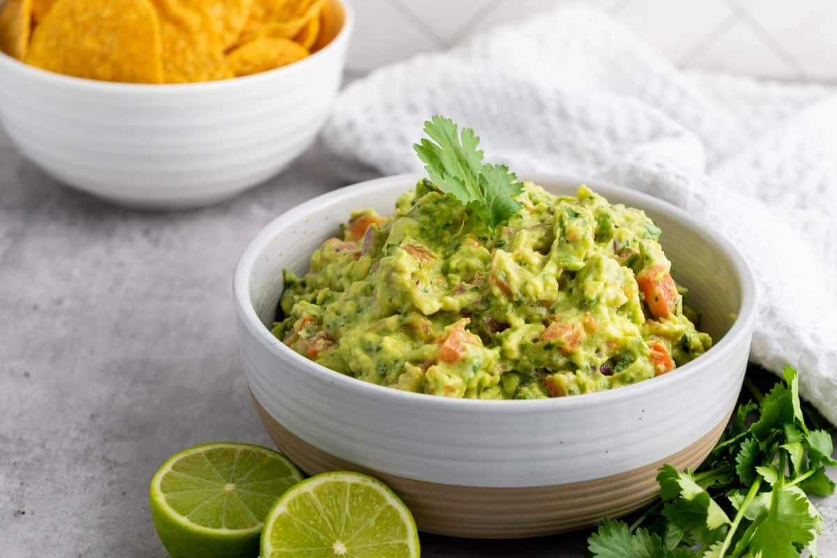 A bowl of guacamole garnished with cilantro sits on a countertop, with lime halves and fresh cilantro nearby. In the background, a bowl of tortilla chips and a white cloth are visible.