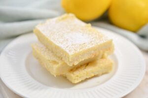 Three lemon bars stacked on a white plate, dusted with powdered sugar, with whole lemons and a light blue cloth in the background.