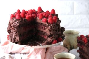 A layered chocolate cake topped and filled with chocolate frosting and fresh raspberries sits on a cake stand. A large slice is missing, revealing the moist, rich interior. Cups of tea and a pink cloth surround the cake.