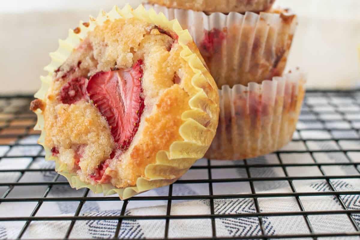 A close-up of strawberry muffins in yellow paper liners, one leaning on a stack of two others, sitting on a black wire cooling rack over a white patterned cloth.