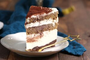 A slice of layered tiramisu cake with cream and cocoa powder sits on a white plate, next to a gold star-shaped fork, on a wooden table with a blue cloth in the background.