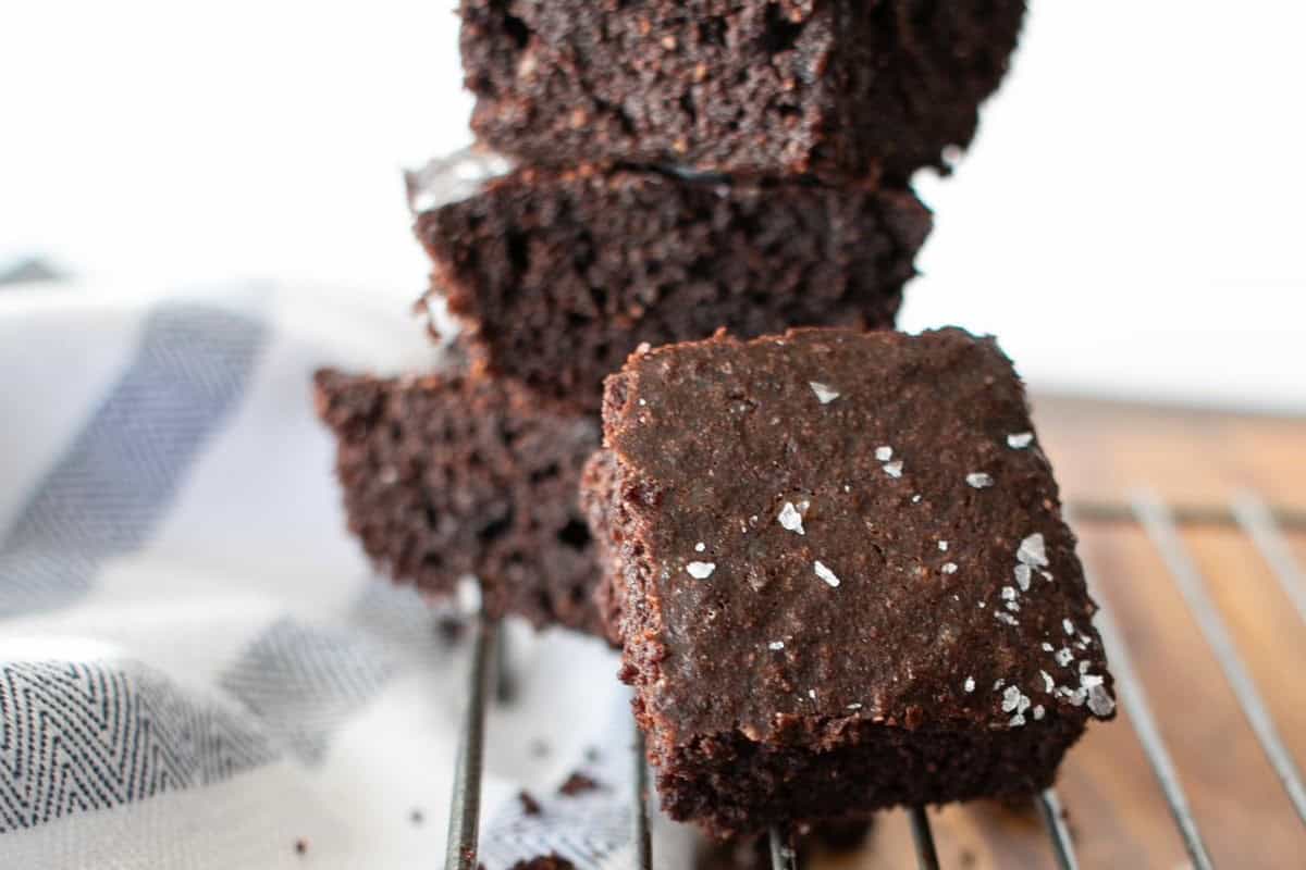 Close-up of rich, fudgy chocolate brownies stacked on a cooling rack, topped with coarse sea salt. A white and blue kitchen towel is partially visible in the background.